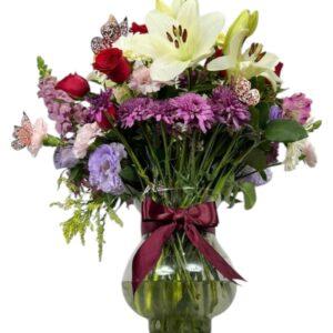 Jarrón de vidrio con lirios blancos, margaritas blancas, rosas rojas, crisantemos morados, lisianthus lila y mariposas decorativas, con lazo color vino.