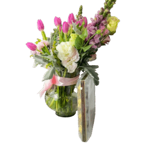 Jarrón de vidrio con tulipanes morados, claveles y crisantemos pastel, follaje gris y lazo rosa, con caja de chocolates Ferrero Rocher.